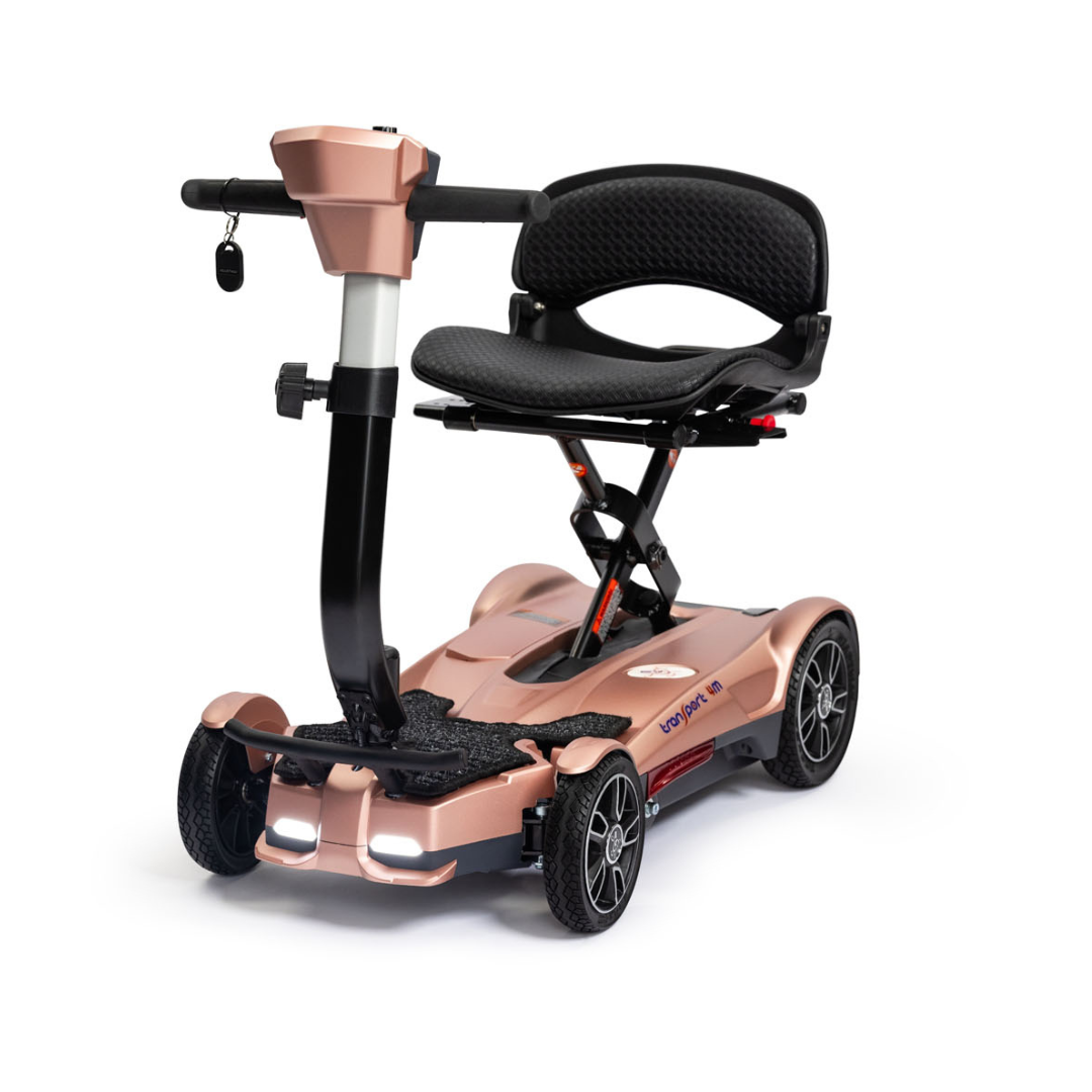The EV Rider Transport 4M Travel Scooter is a compact, rose-gold, four-wheel scooter with a black cushioned seat, armrests, handlebars, ignition key, and an elevated folding seat for convenient travel.